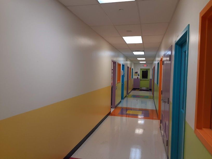 Colorful school hallway with white walls, orange stripe, blue doors, and fluorescent ceiling lights.