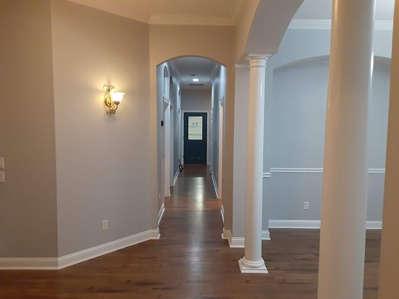 Empty hallway with hardwood floors, beige walls, white columns, and a wall sconce light