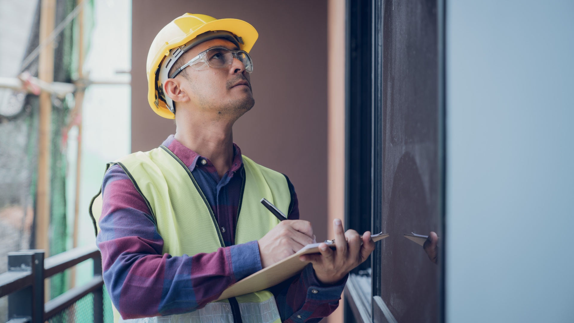 Worker in yellow hard hat and vest using a clipboard beside a building window