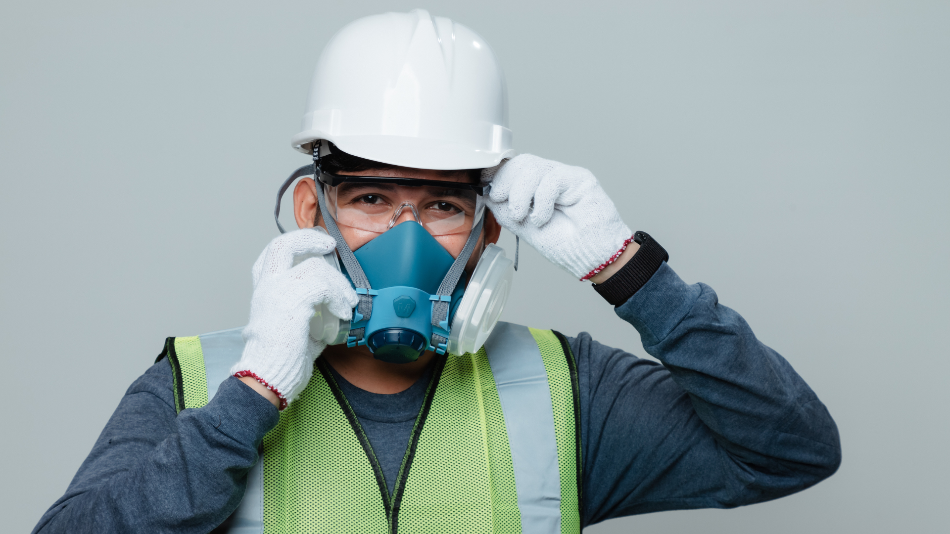 Worker in white hard hat adjusting a blue respirator mask, wearing gloves and a reflective vest.