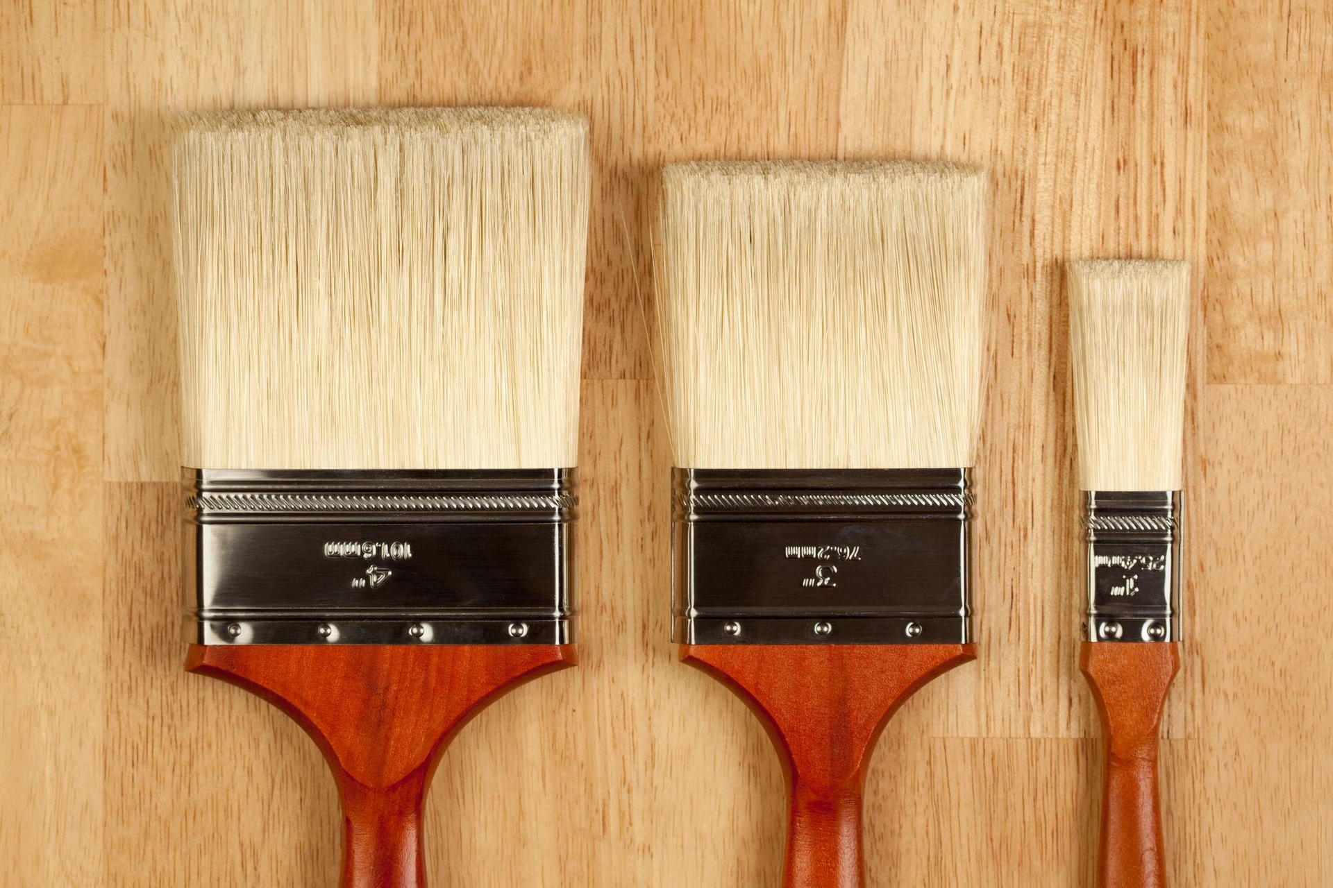 Three paintbrushes with wooden handles lined up on a wooden surface, largest to smallest.