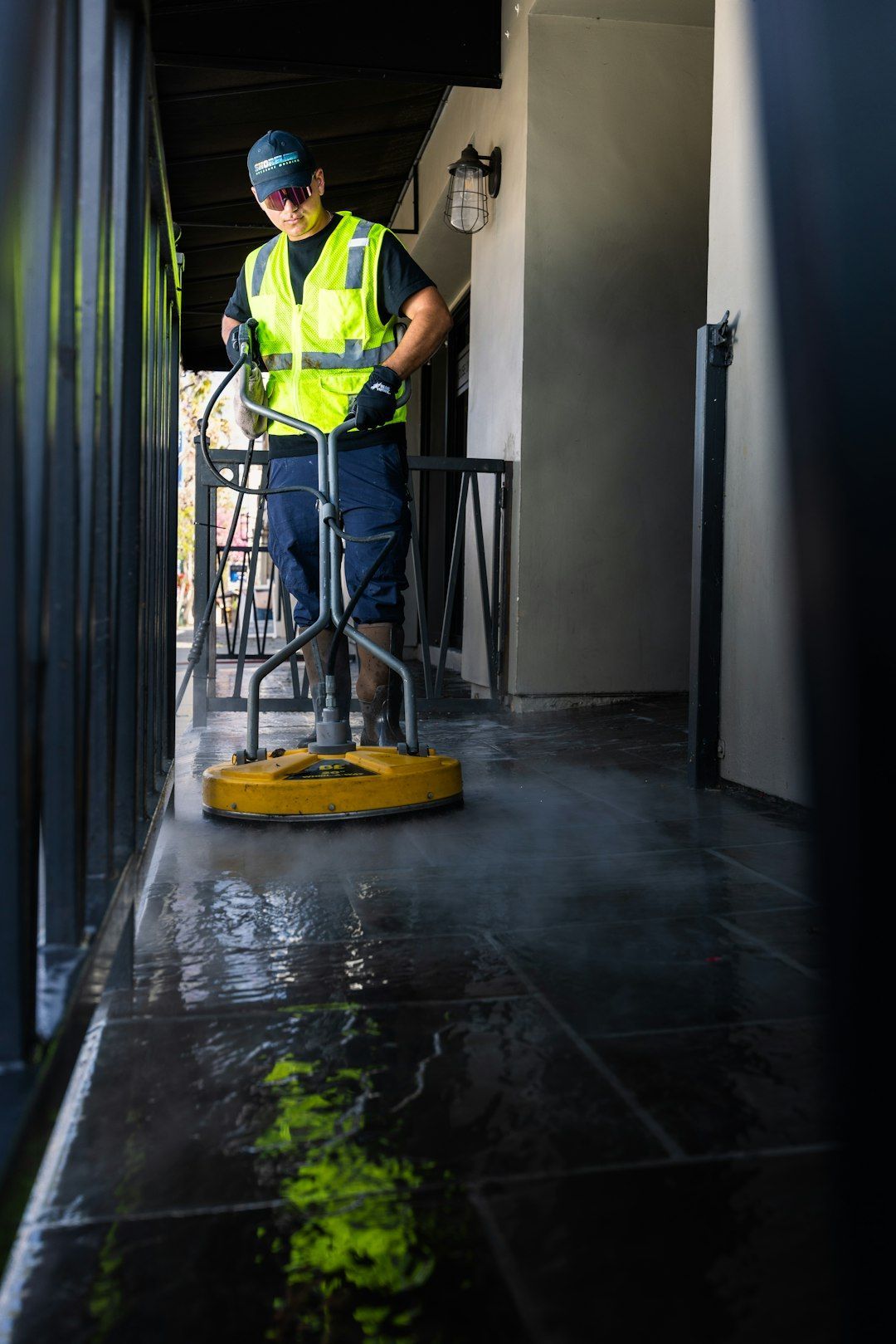 Worker in a high-visibility vest cleaning a wet hallway floor with a floor scrubber.