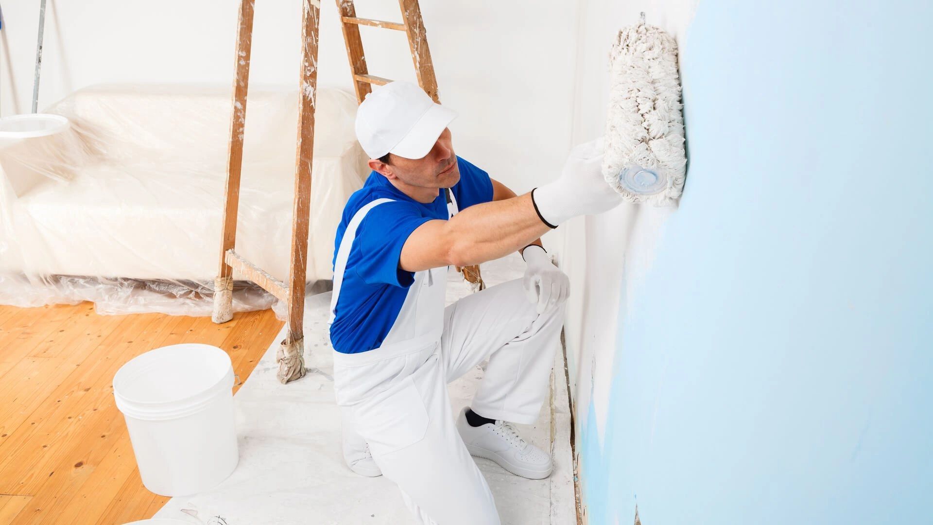 Painter applying white paint to a light blue wall in a bright room with a ladder and paint bucket.