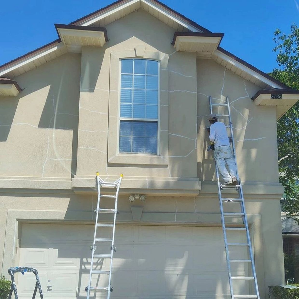 Worker painting exterior of beige two-story house while standing on a ladder