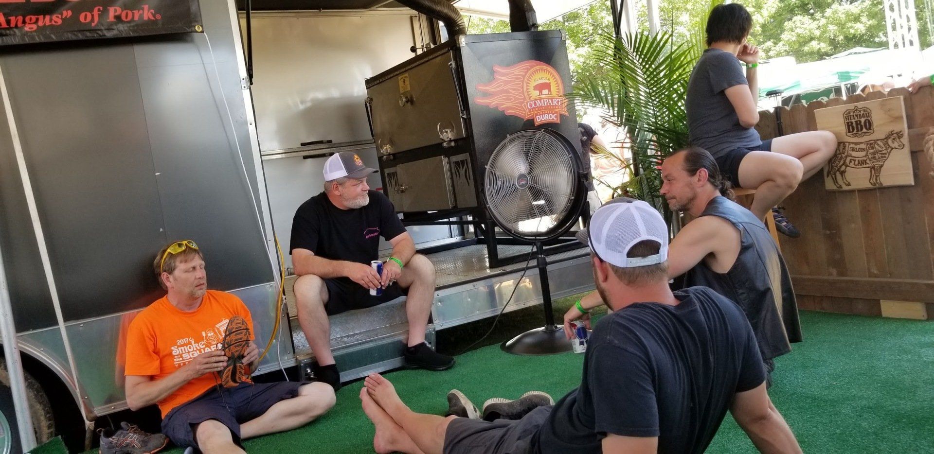 A group of men are sitting on the grass in front of a food truck.