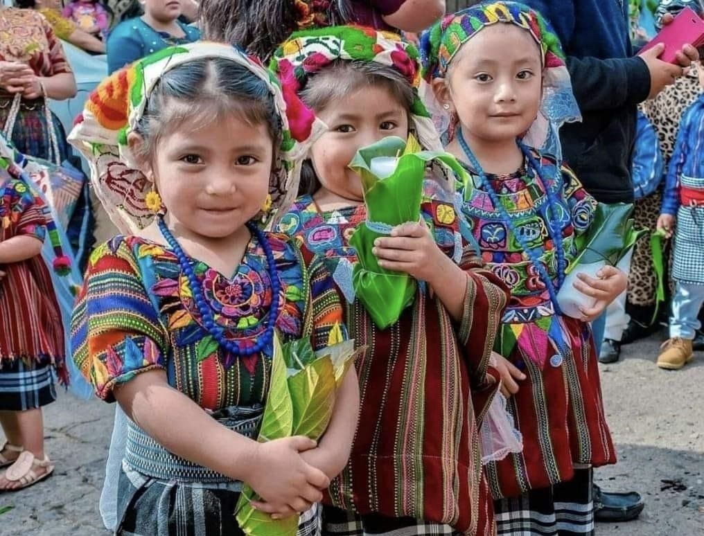 Drei Mädchen in farbenfroher traditioneller guatemaltekischer Kleidung, lächelnd.