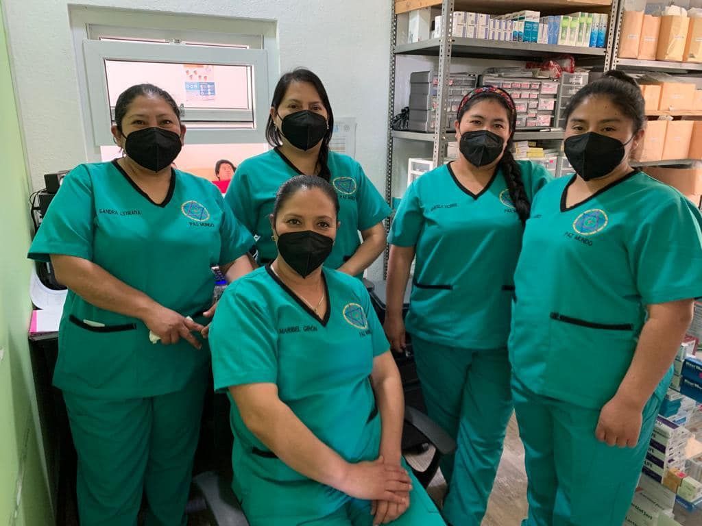 Fünf Frauen in türkisfarbenen OP-Kitteln und mit Masken in einer Apotheke, lächelnd und in die Kamera schauend.