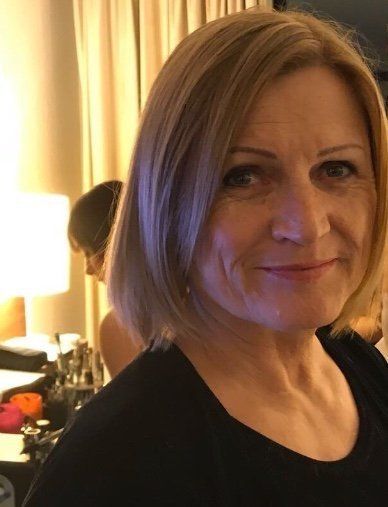 Frau mit kurzen blonden Haaren lächelt, sie trägt ein schwarzes Oberteil. Innenraum, sanftes Licht.