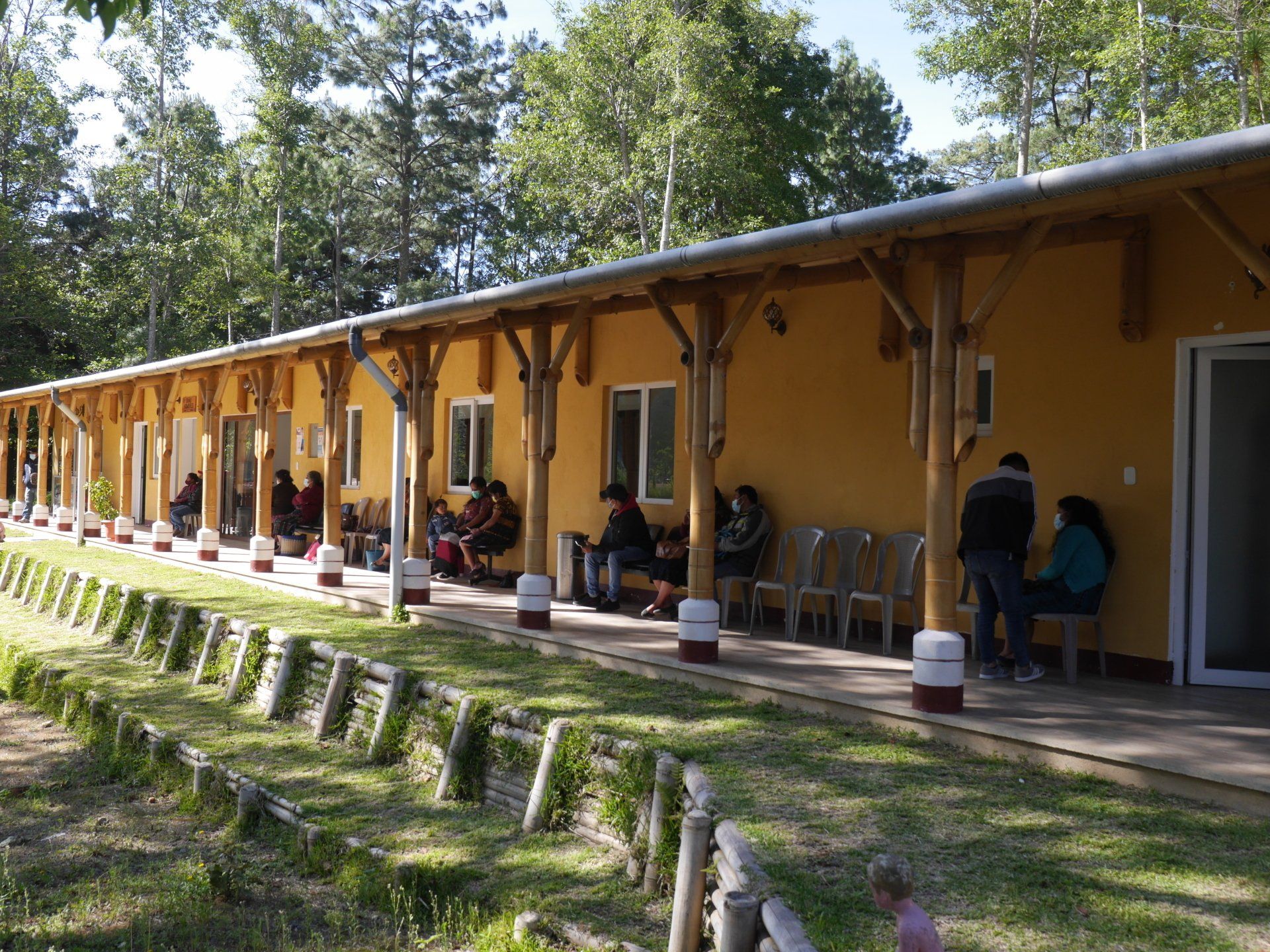 Gelbes Gebäude mit überdachter Veranda; Menschen sitzen draußen. Im Hintergrund grünes Gras und Bäume.