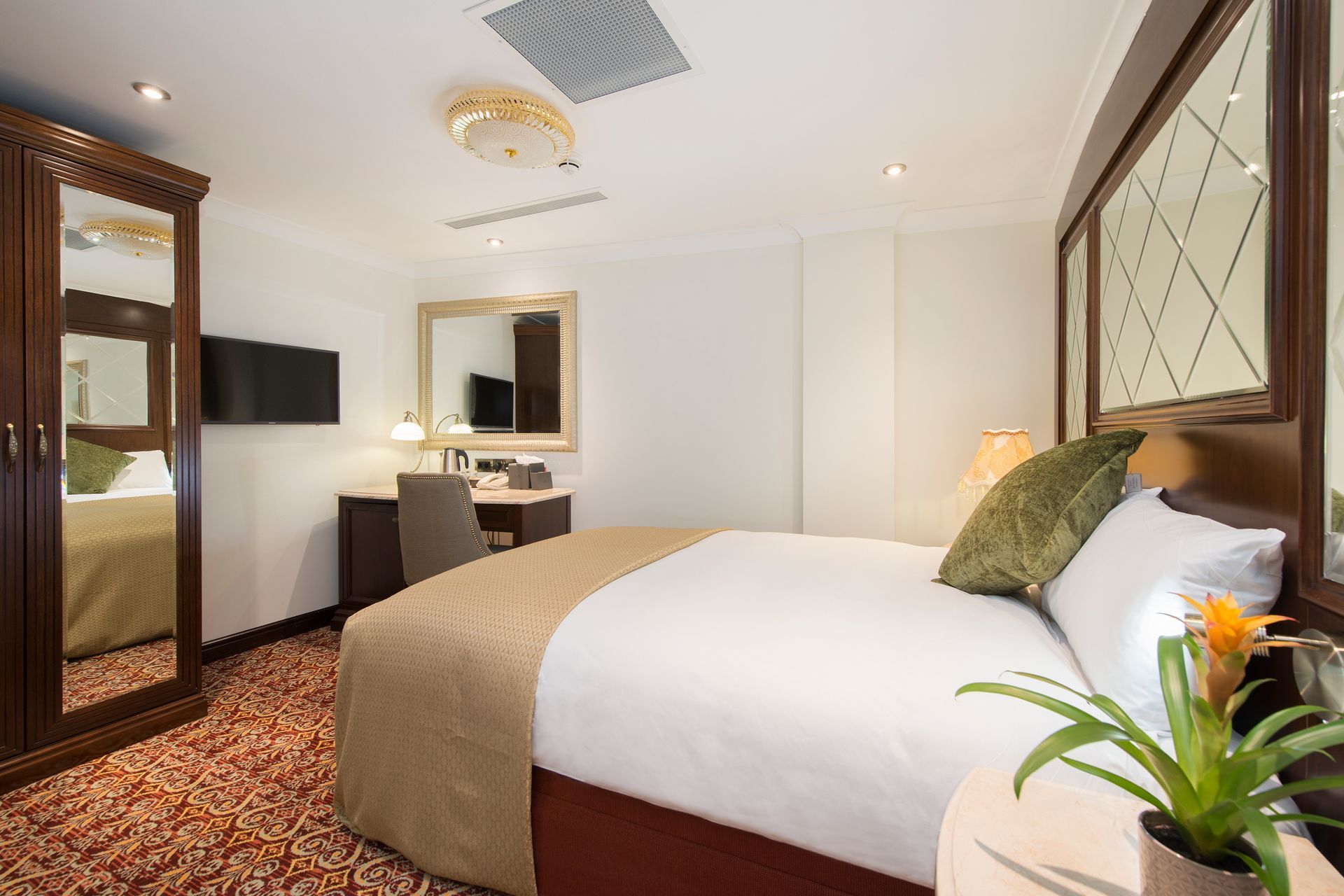 Hotel room with bed, desk, mirror, and wardrobe, with patterned carpet and neutral colors.