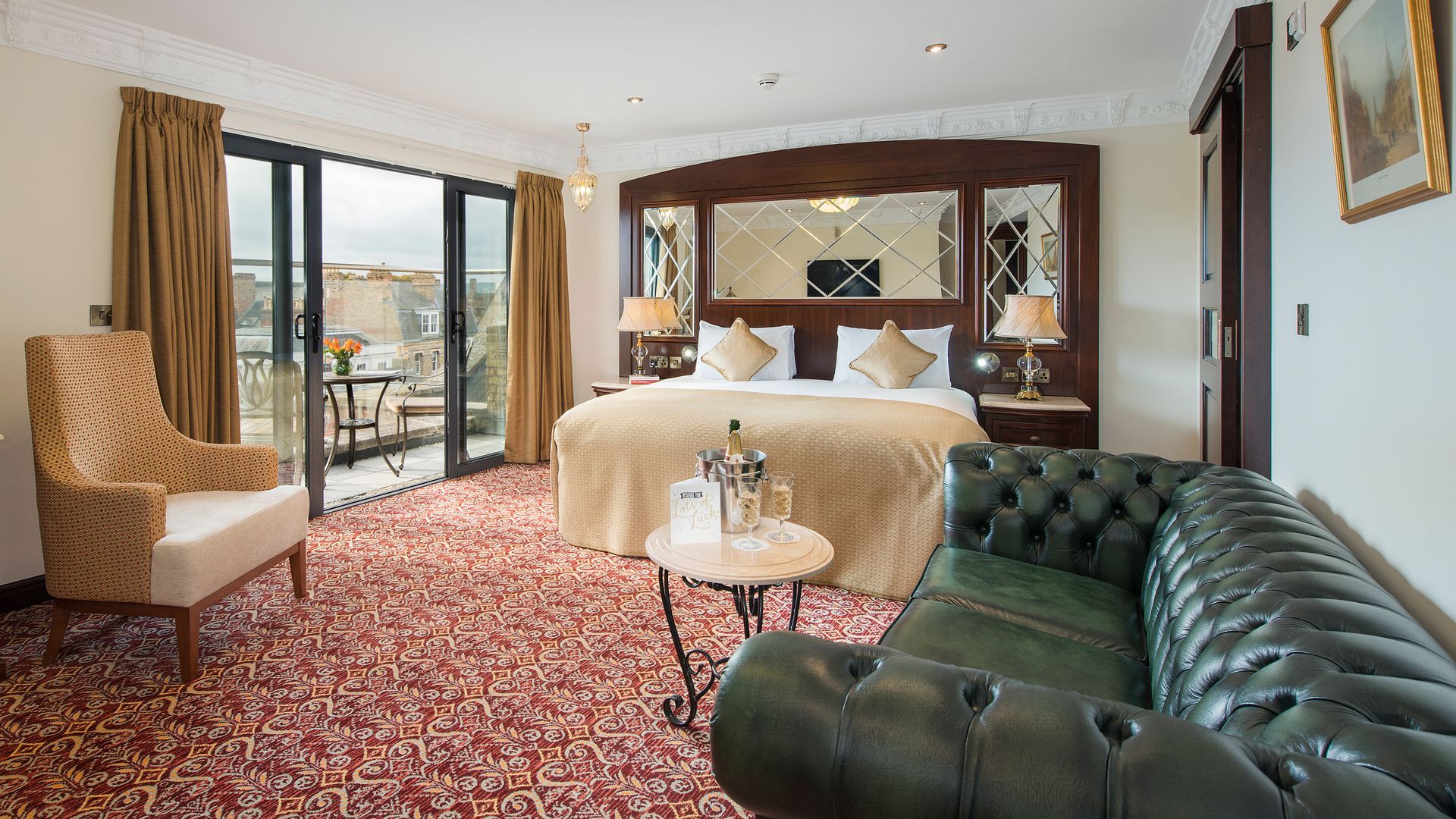 Luxurious hotel room with bed, sofa, balcony access, and red patterned carpet.