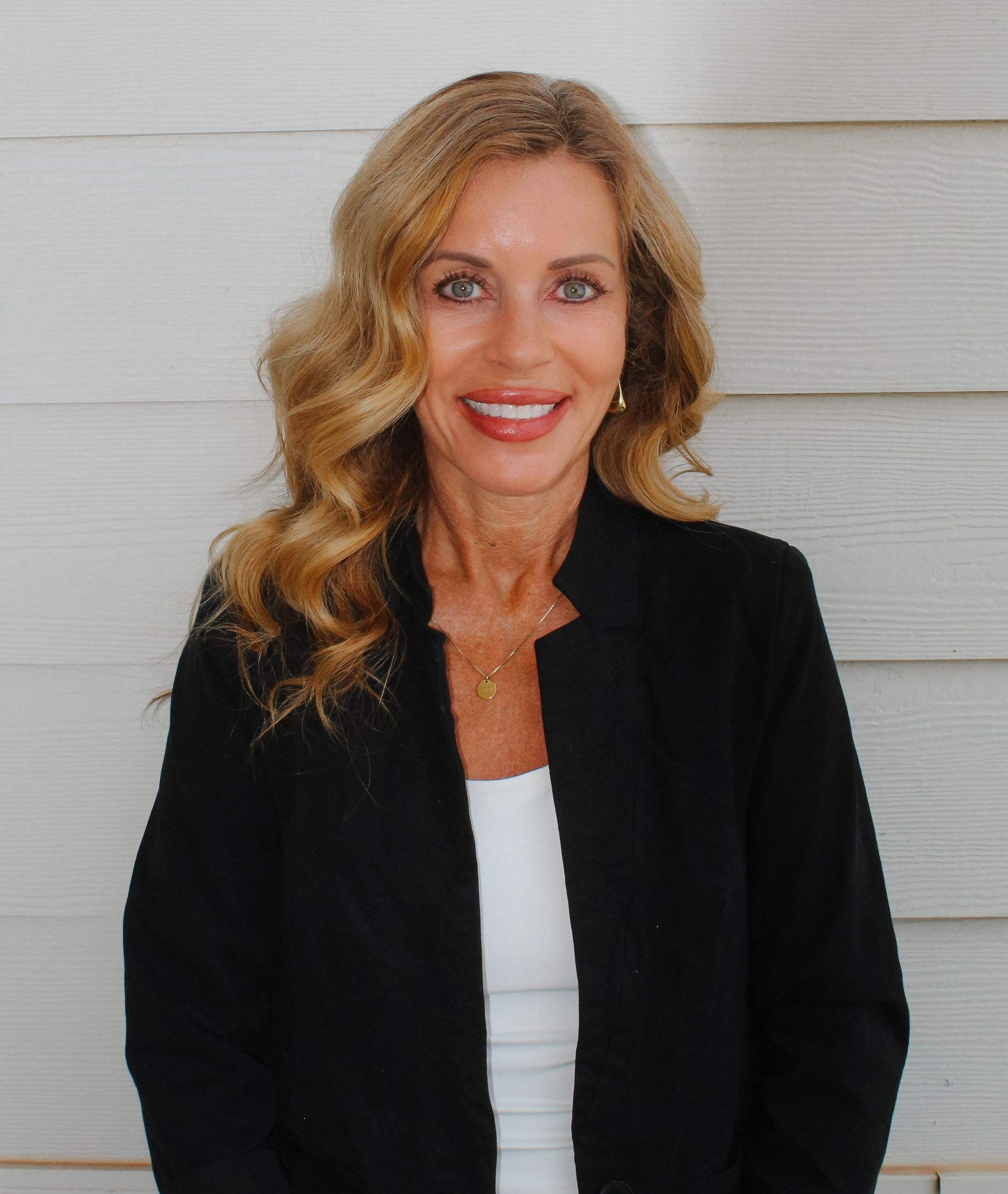 A person with long dark hair smiles while sitting on a white couch, wearing a black shirt, light jeans, and sandals.