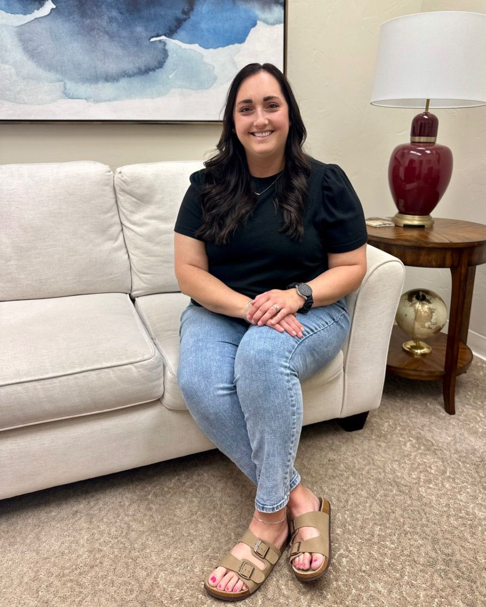 A person with long dark hair smiles while sitting on a white couch, wearing a black shirt, light jeans, and sandals.