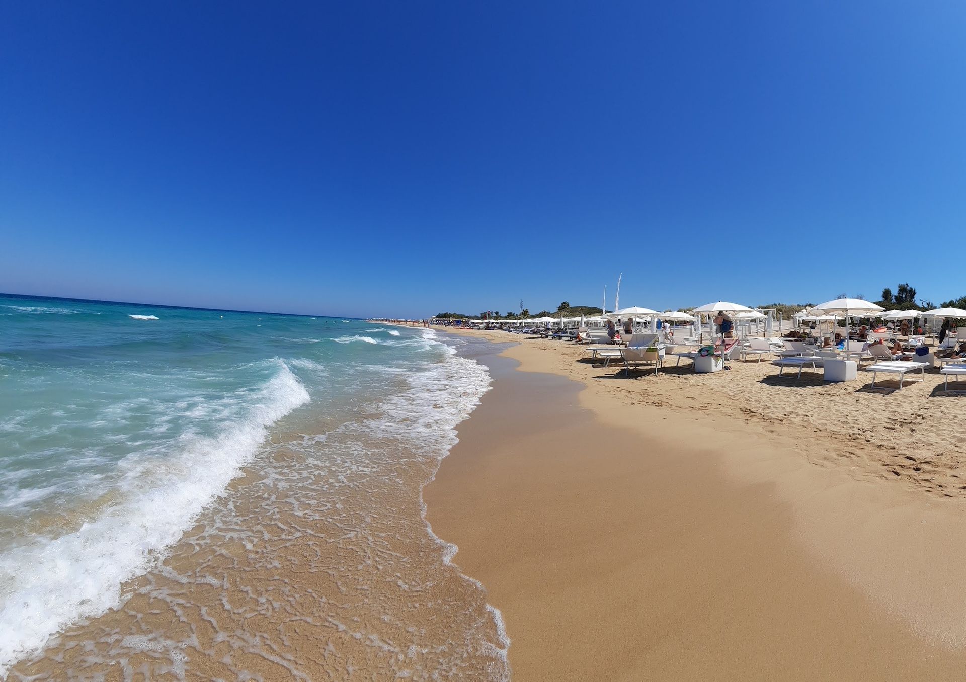 Monopoli, Capitolo Spiaggia sabbiosa con onde turchesi sotto un cielo azzurro e limpido. Ombrelloni bianchi e lettini prendisole costeggiano la riva.