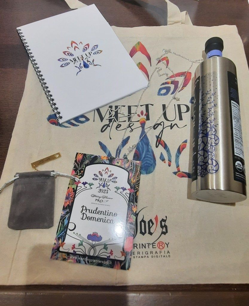 Borsa tote con blocco note, bottiglia d'acqua, piccola custodia e biglietto; il tutto con un disegno floreale su una superficie in legno.