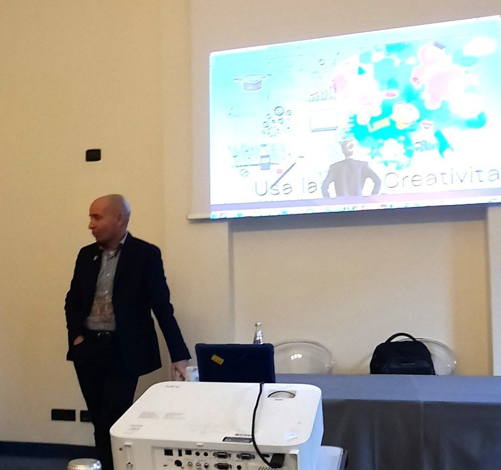 Un uomo in giacca e cravatta tiene una presentazione, in piedi accanto a un tavolo con un proiettore e uno schermo sullo sfondo.