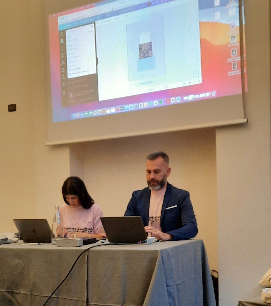 Due persone sedute a un tavolo con i loro computer portatili, una presentazione sullo schermo in alto.