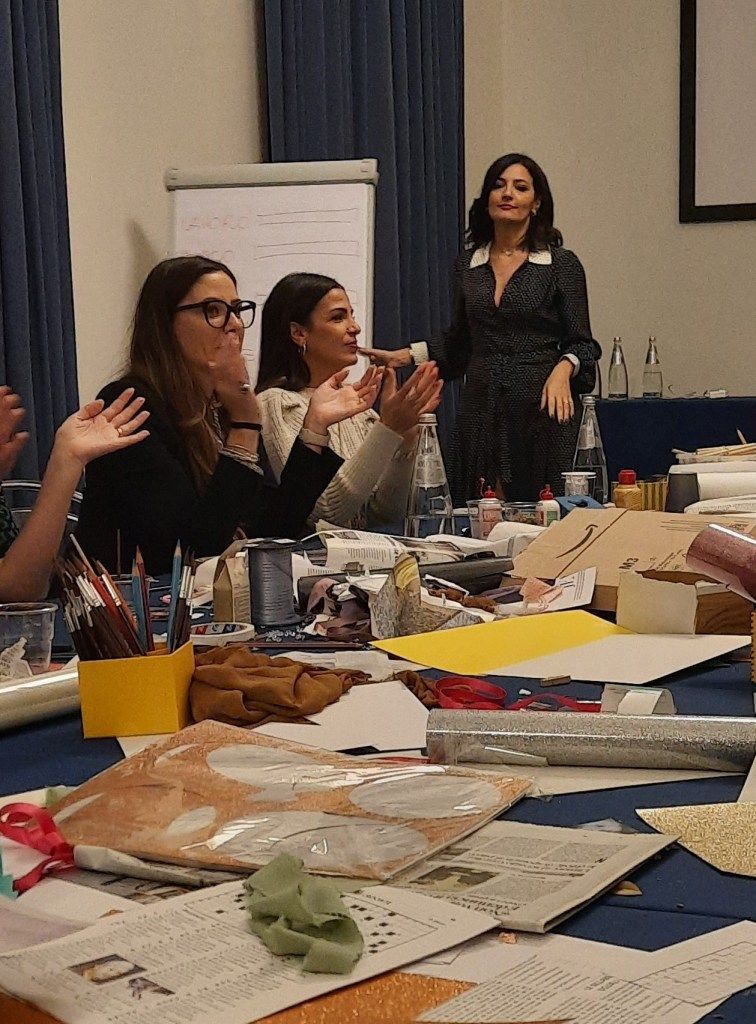 I partecipanti al workshop applaudono, il relatore in abito a pois fa gesti. Tavolo con oggetti artigianali e materiali.