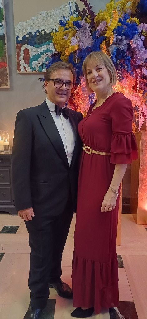 Una coppia in abito formale in piedi vicino a una colorata composizione floreale. La donna indossa un abito bordeaux con una cintura dorata.