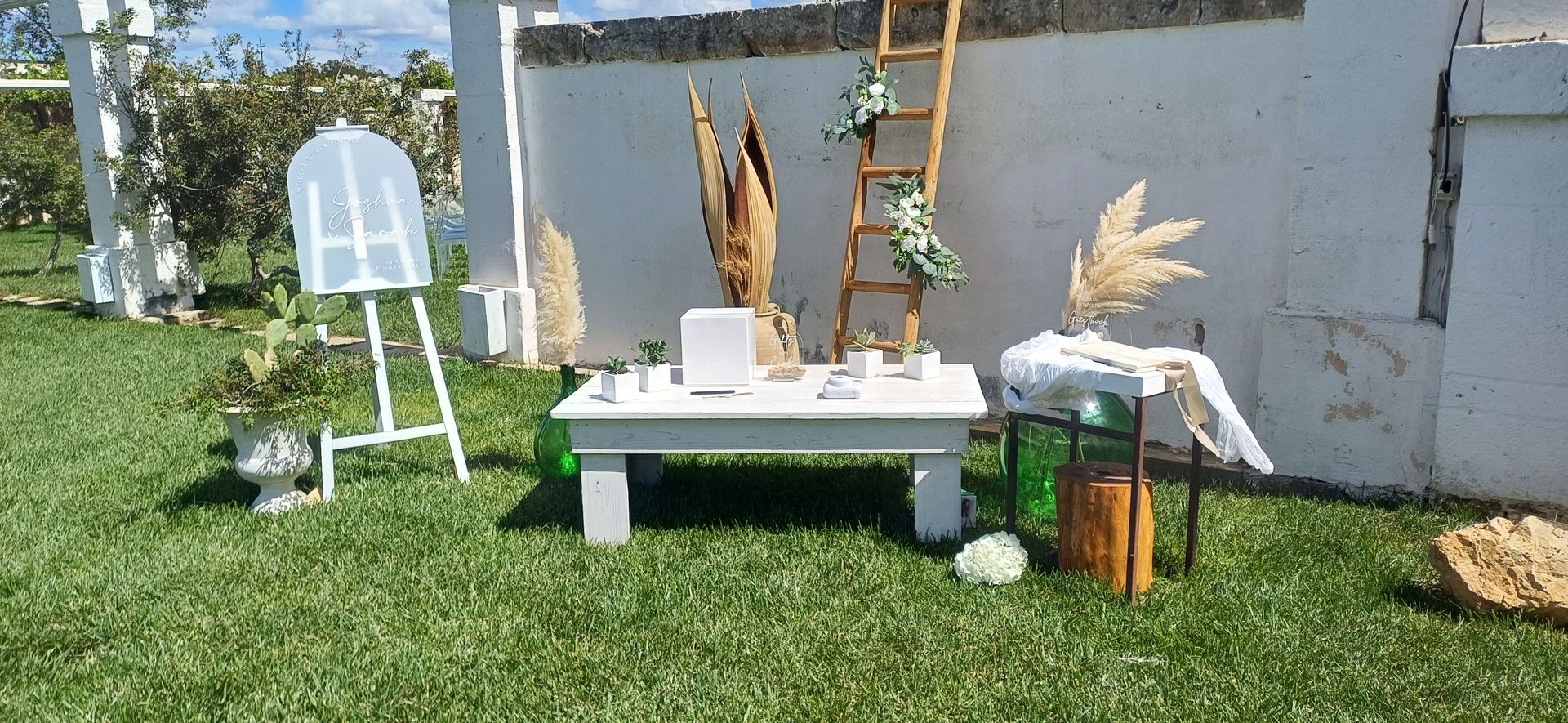 Decorazioni per matrimonio su prato verde con tavolo bianco, cavalletto e scala contro un muro bianco.