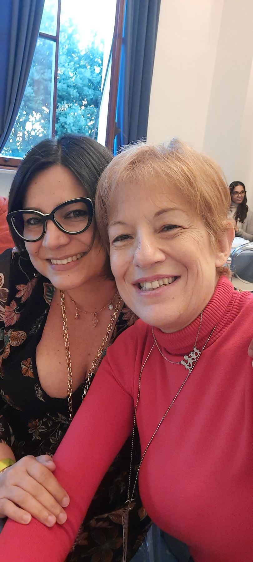 Due donne sorridenti si scattano un selfie. Una indossa occhiali, top nero e una lunga collana. L'altra indossa un dolcevita rosso.