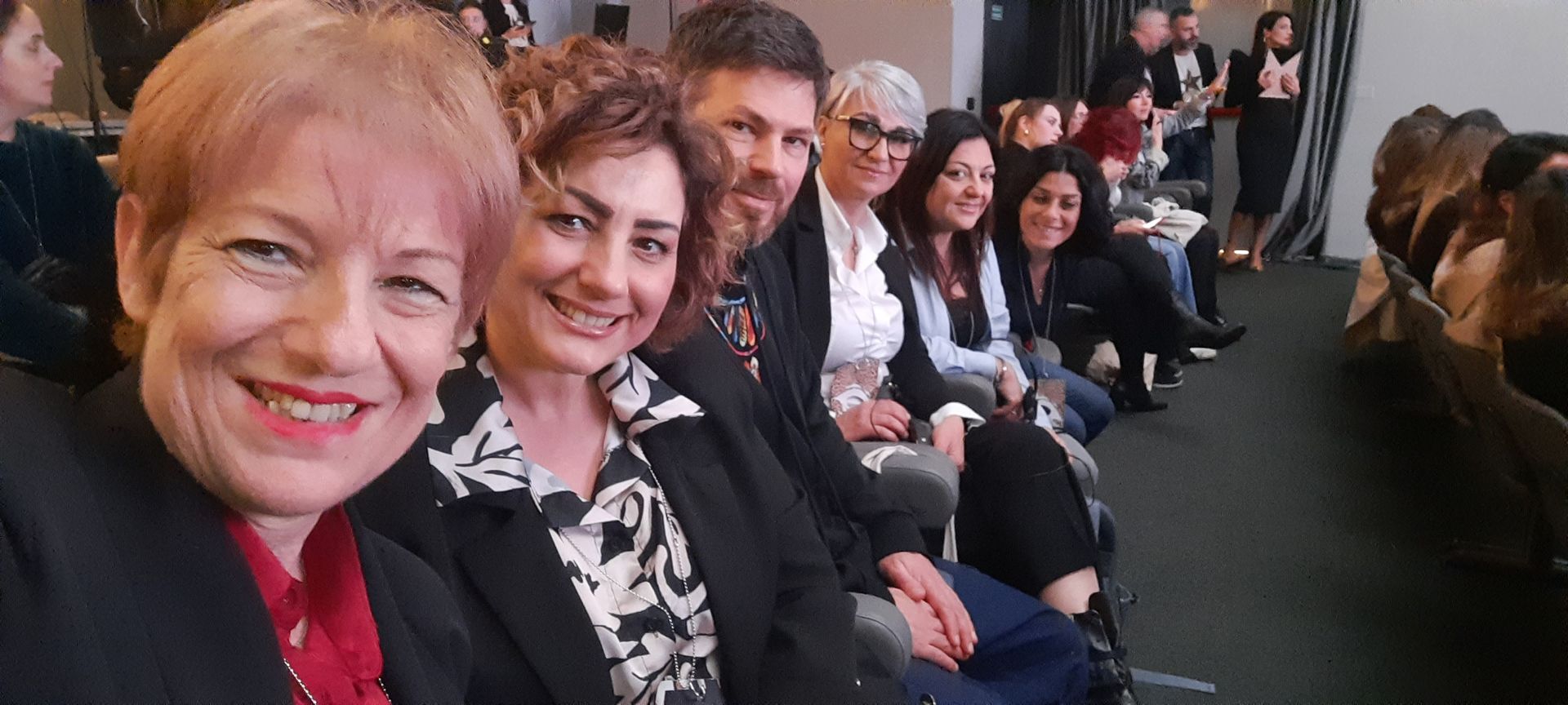 Un gruppo di persone sorridenti è seduto su una fila di sedie. Alcune guardano verso la telecamera. All'interno, luce del giorno.
