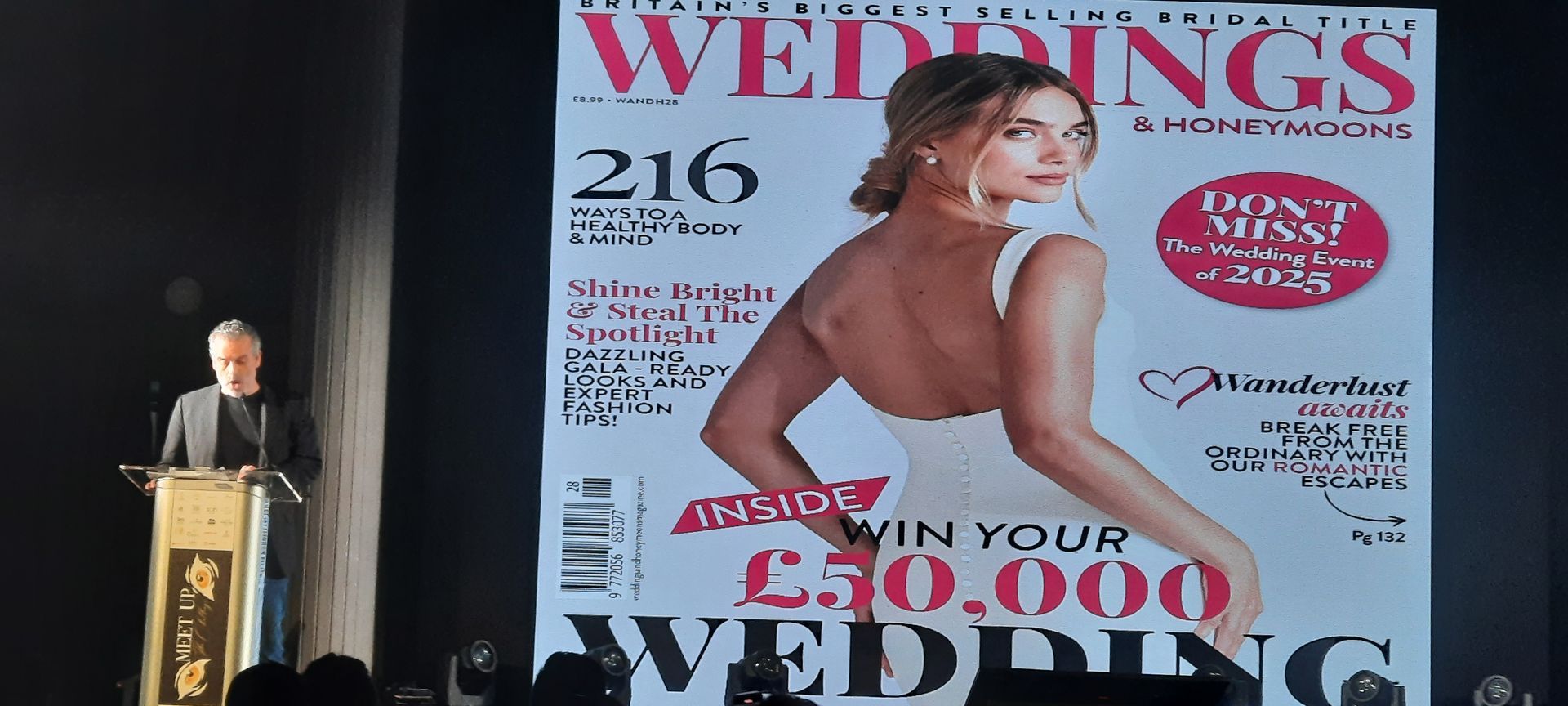 Un uomo parla su un podio accanto alla copertina di una grande rivista insieme a una donna in abito da sposa.