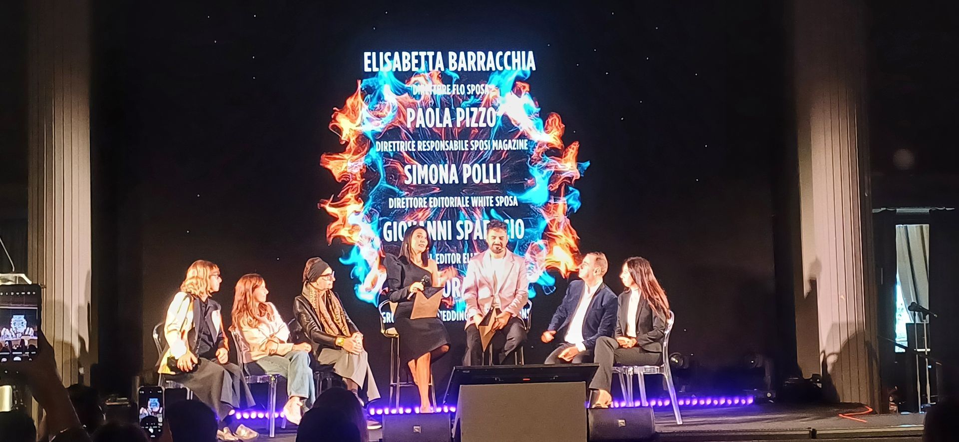 Un gruppo di persone su un palco con uno schermo su cui sono visualizzati i nomi, circondato da fiamme.