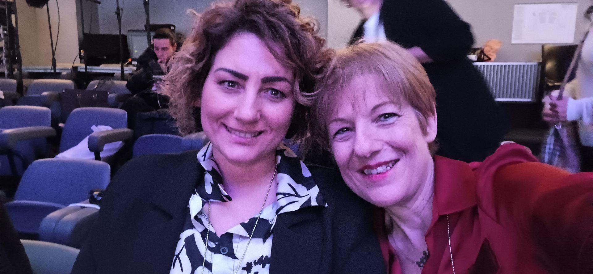 Due donne sorridenti si scattano un selfie in un auditorium. Una indossa una giacca nera e una camicia a fantasia, l'altra un top rosso.