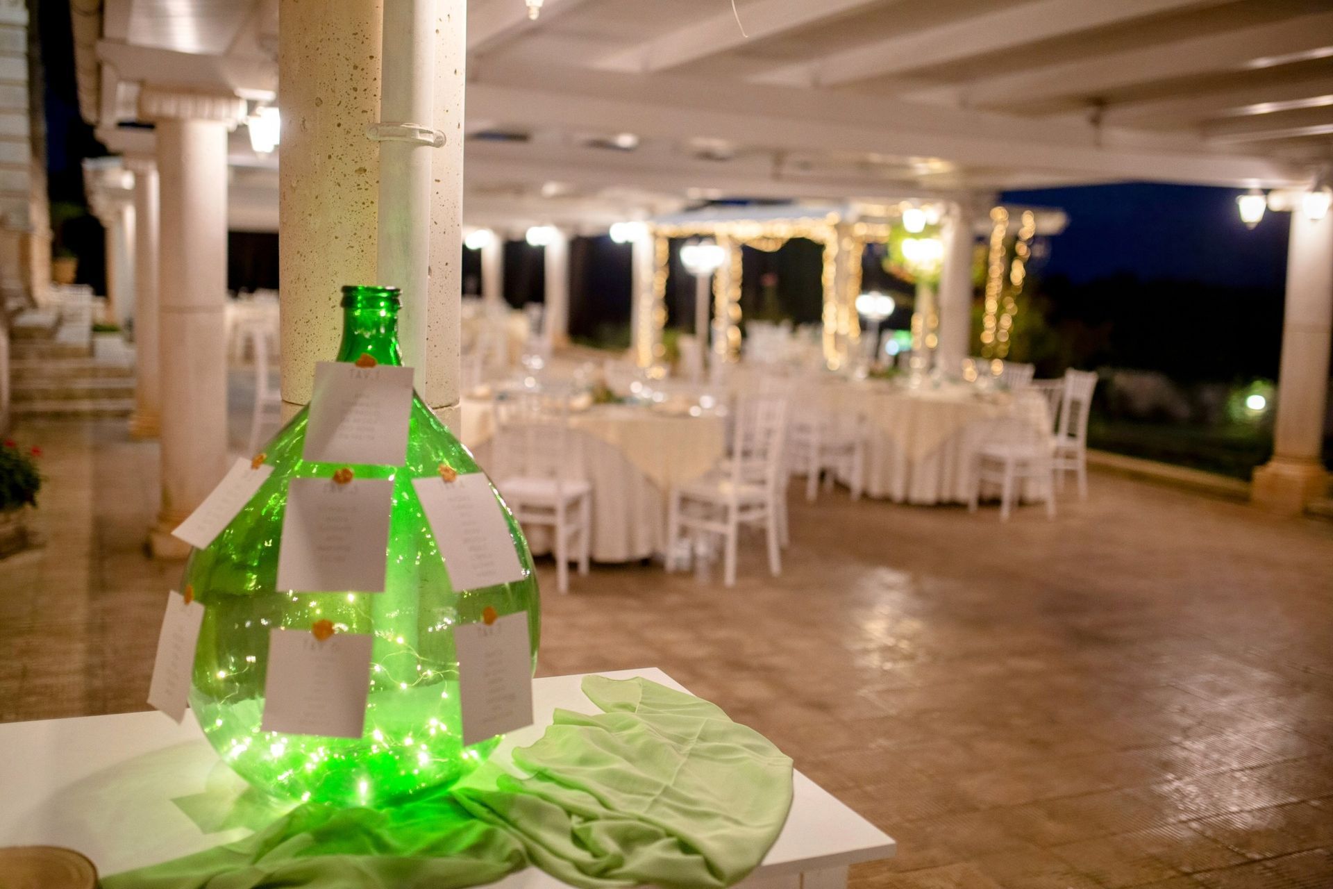 Espositore luminoso a forma di bottiglia verde con segnaposto, davanti ai tavoli apparecchiati per un evento formale all'aperto.