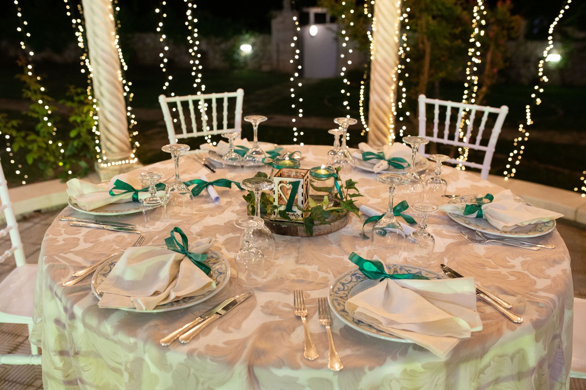 Tavolo rotondo apparecchiato per una cena formale, decorato con decorazioni verdi e bianche, illuminato da luci a stringa.