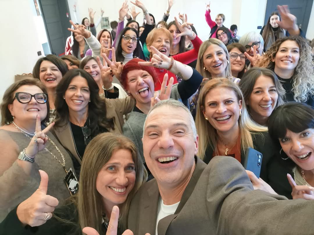 Un gruppo di persone sorridenti posa per un selfie. Molti alzano le mani e fanno il segno della pace. Al chiuso.
