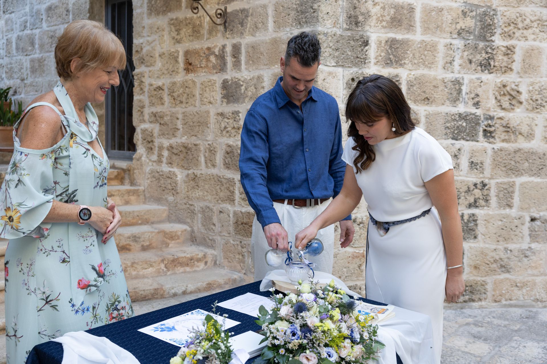 Una coppia versa della sabbia in un bicchiere durante una cerimonia nuziale all'aperto, alla quale partecipa una donna con un abito floreale.