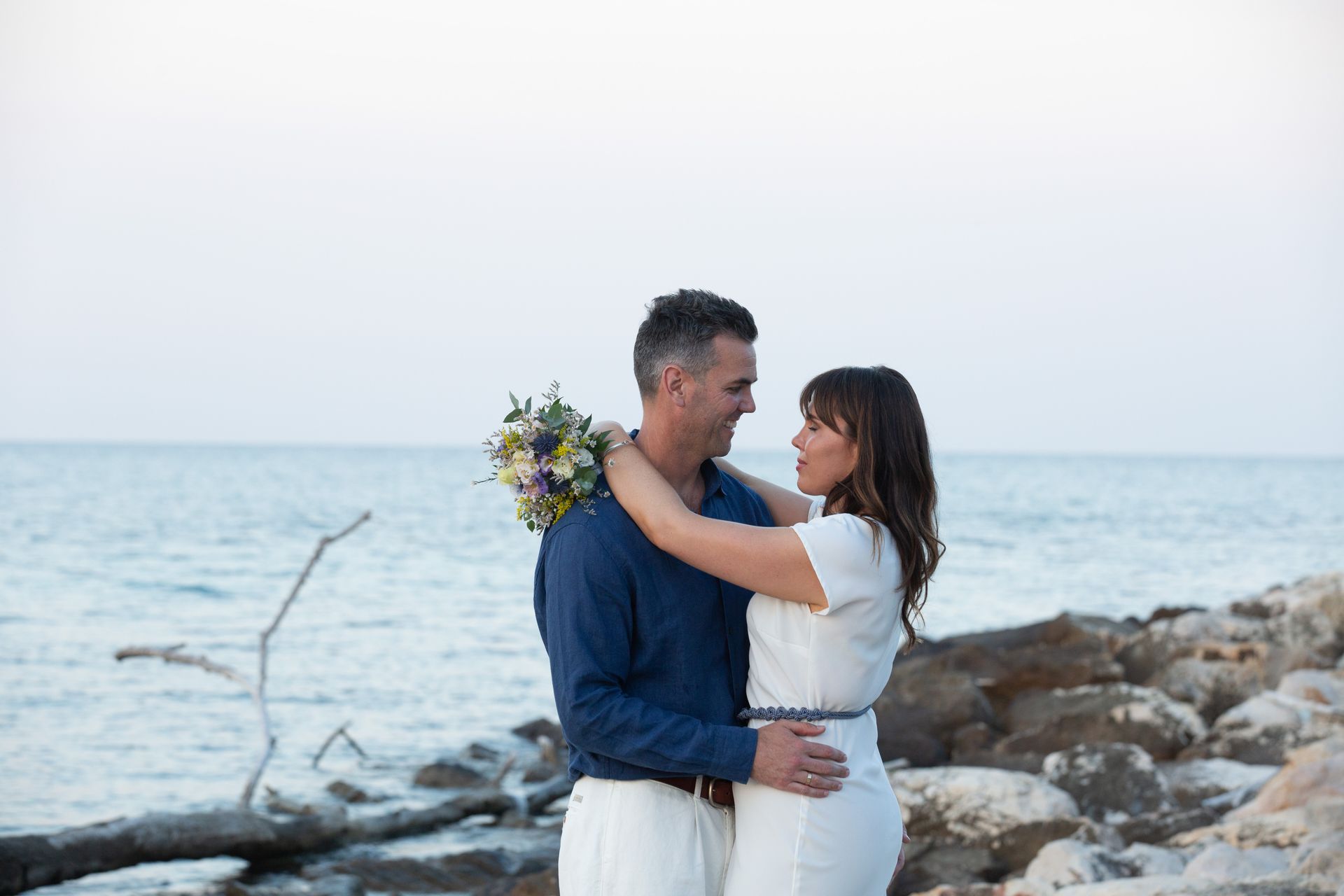 Coppia abbracciata su una spiaggia rocciosa, che si guarda. Uomo in blu, donna in abito bianco, con in mano un bouquet. Sullo sfondo l'oceano.
