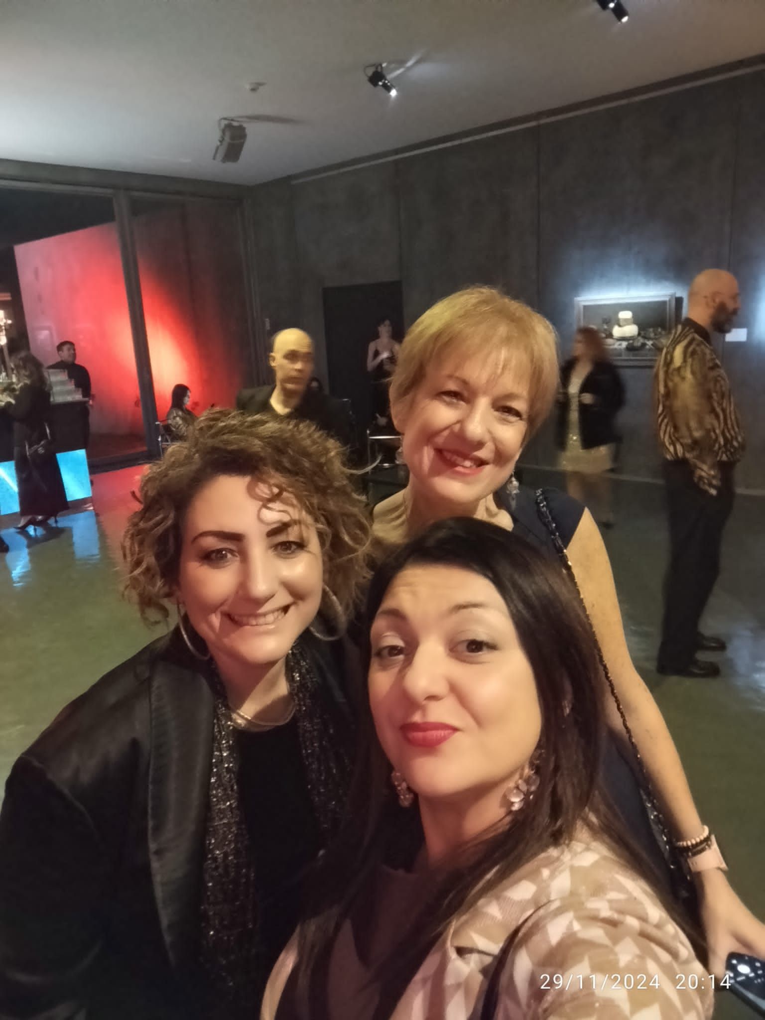 Tre donne sorridono per un selfie a un evento. Sullo sfondo, altre persone e luci rosse.