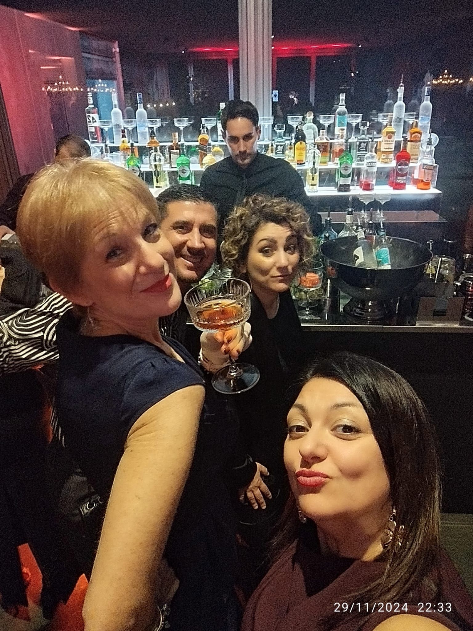 Gruppo di persone sorridenti in posa davanti a un bar, con un barista sullo sfondo.
