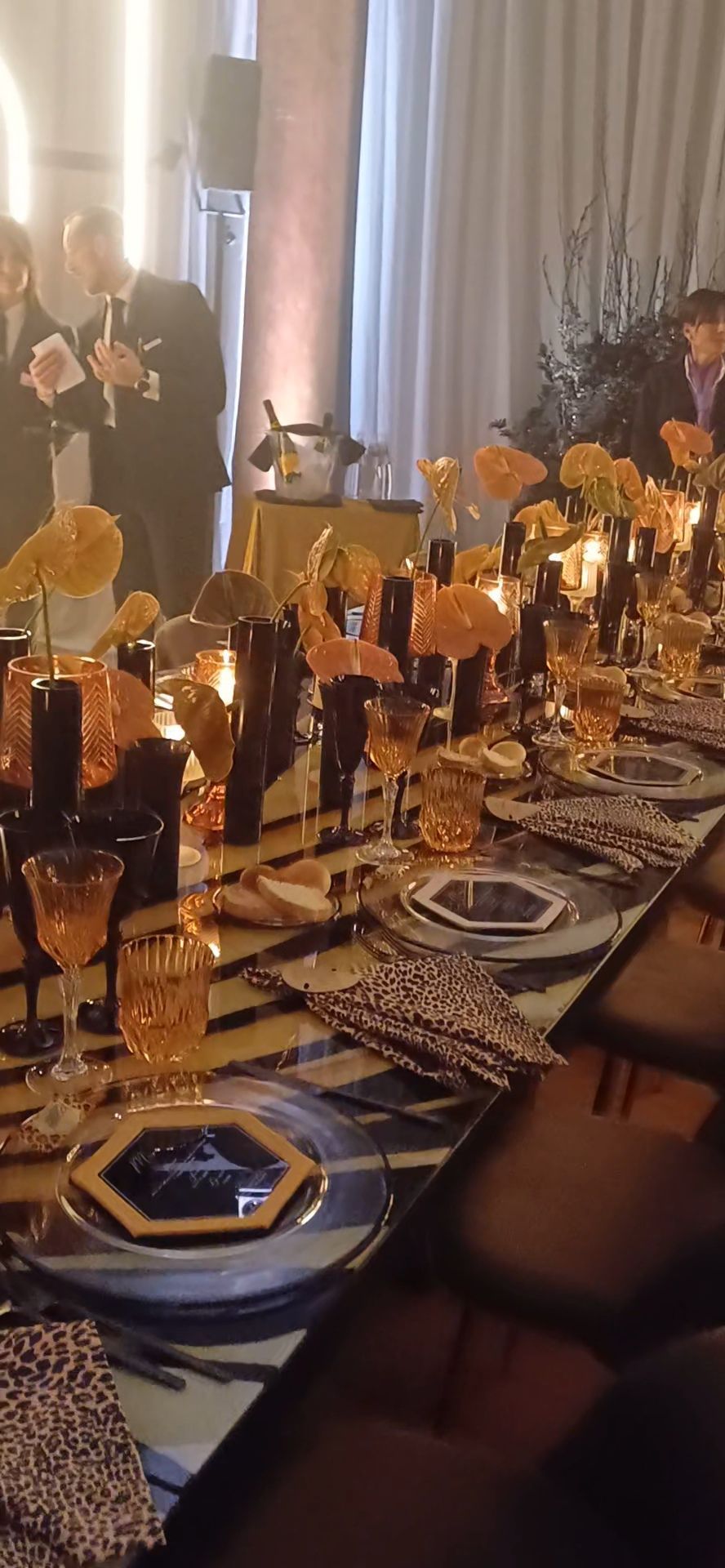 Tavolo da pranzo formale apparecchiato con decorazioni arancioni e nere, ospiti sullo sfondo.