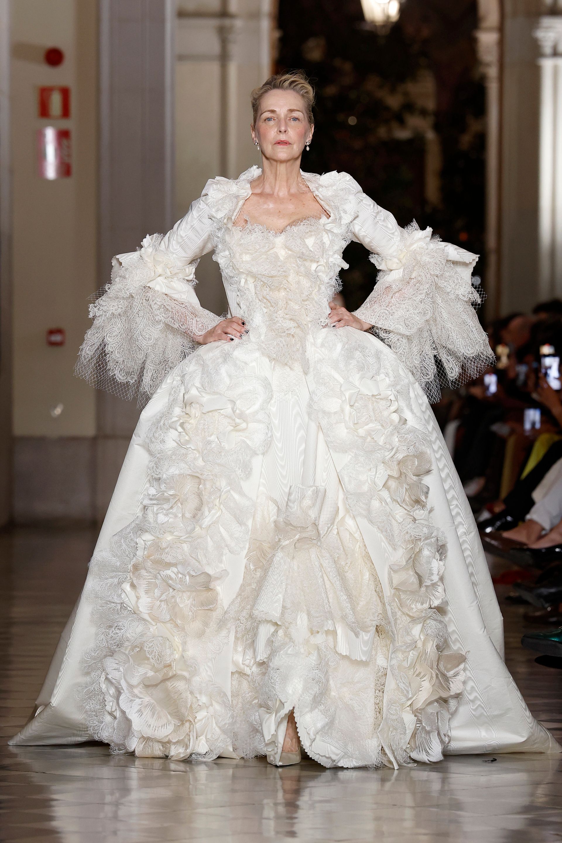 Donna in un elaborato abito bianco con ampia gonna, maniche a balze e decorazioni floreali, su una passerella.