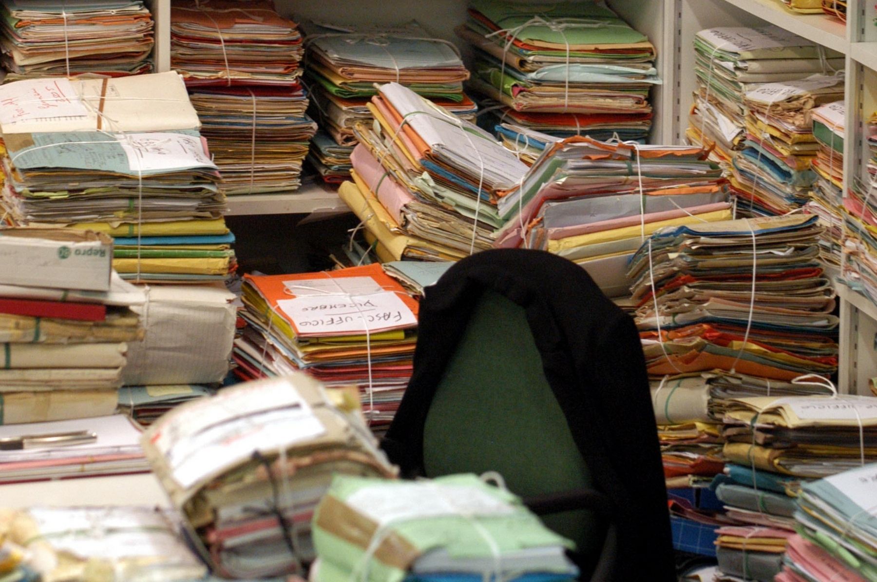 Pile di documenti cartacei legati ingombrano uno spazio di lavoro con una sedia al centro.