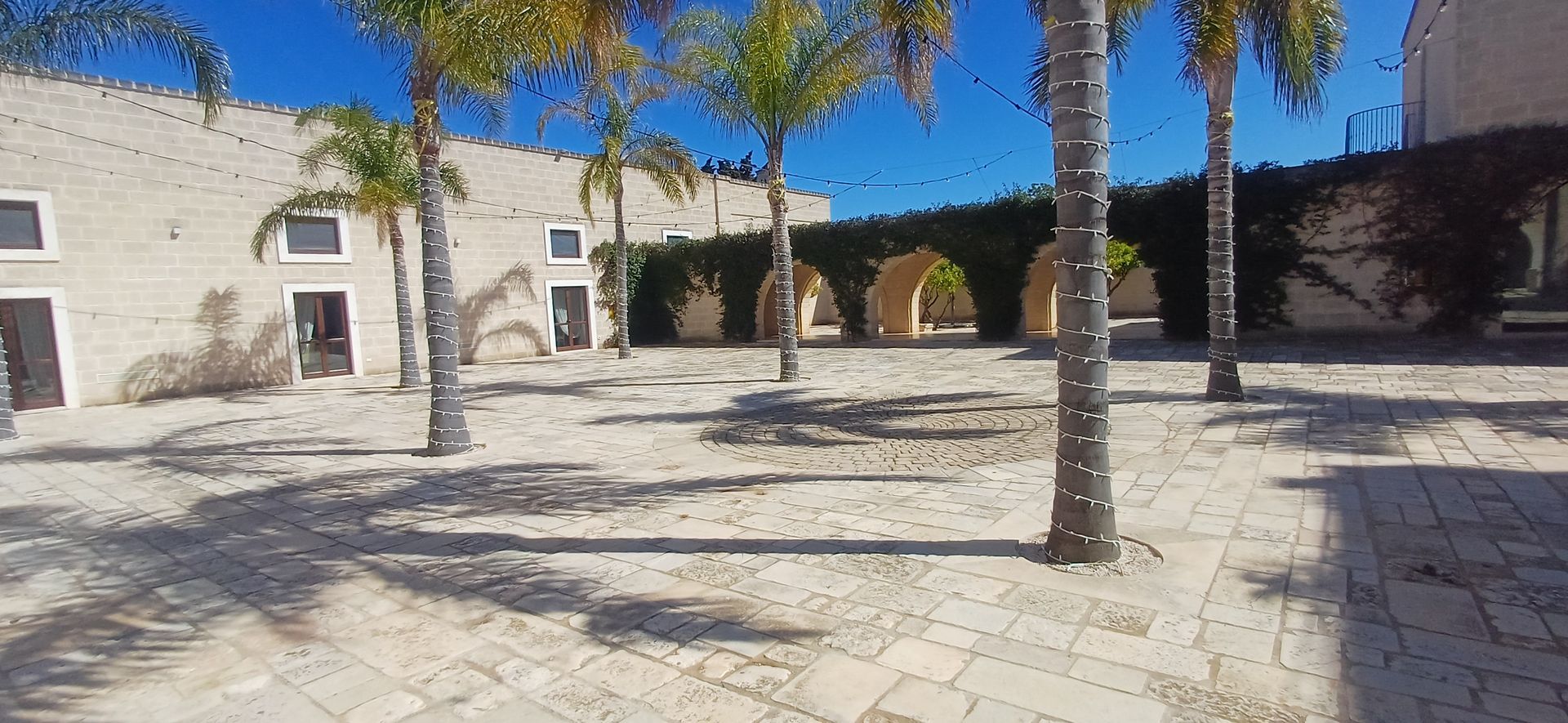 Palme in un cortile con pavimento in pietra chiara e un edificio con ingressi ad arco.