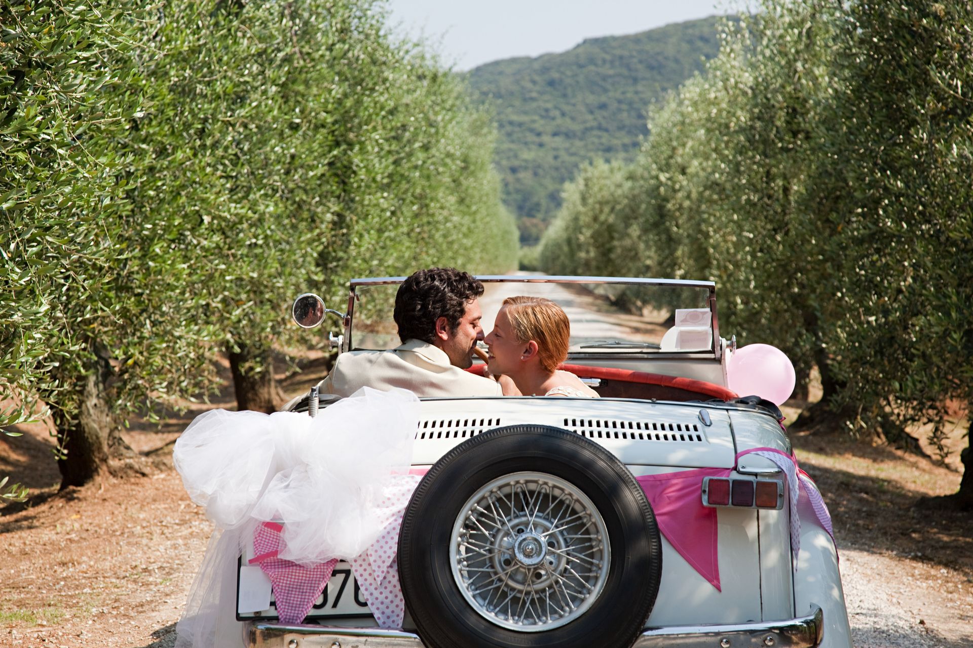 Una coppia si bacia in un'auto d'epoca decorata per un matrimonio, mentre attraversa un uliveto.