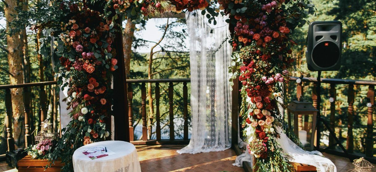 Arco nuziale decorato con fiori rossi, con vista sulla foresta. Un oratore è in piedi nelle vicinanze.