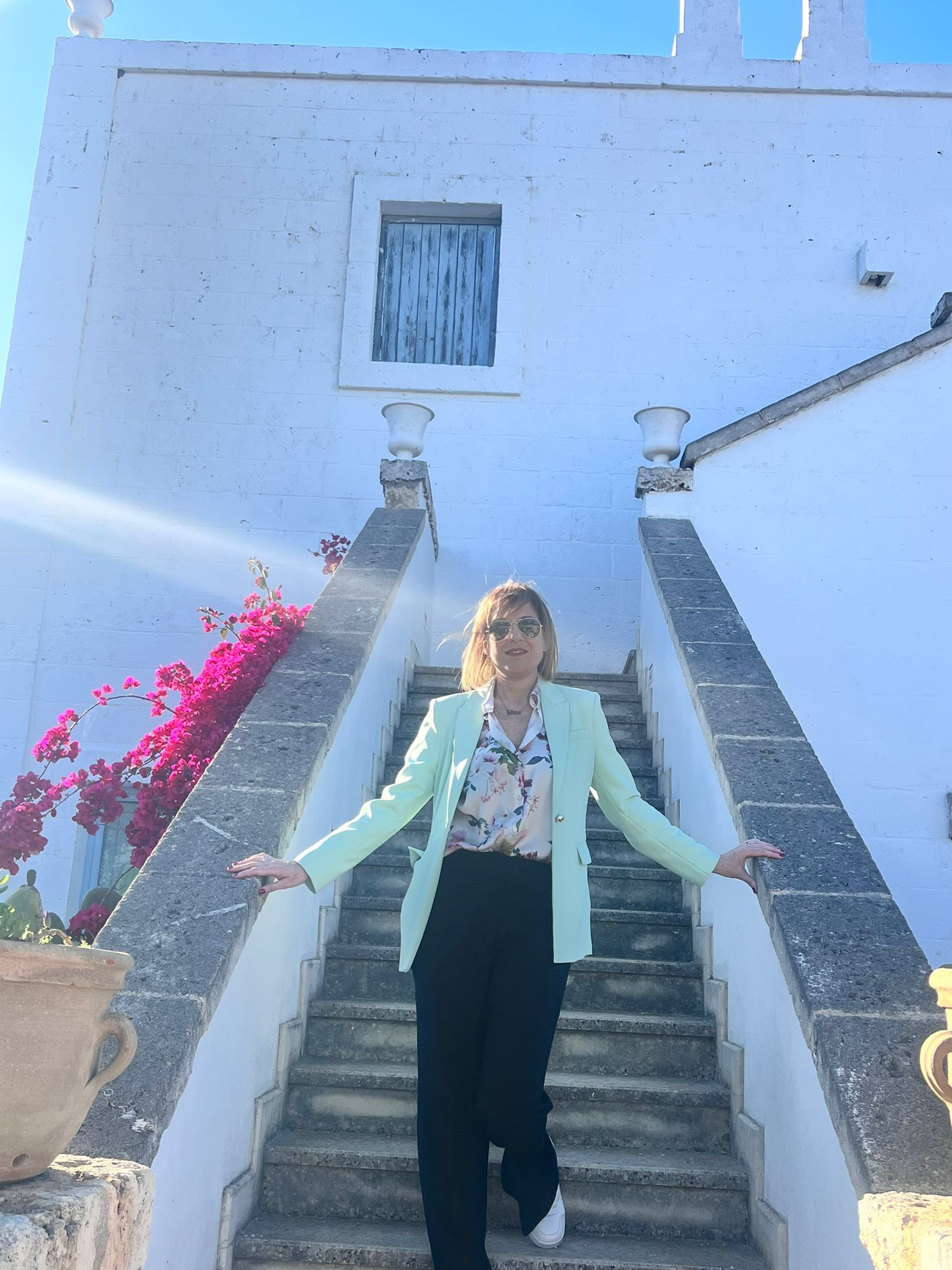 Una donna con una giacca verde chiaro è in piedi su una scala bianca, con le braccia tese. Un edificio bianco, un cielo luminoso.
