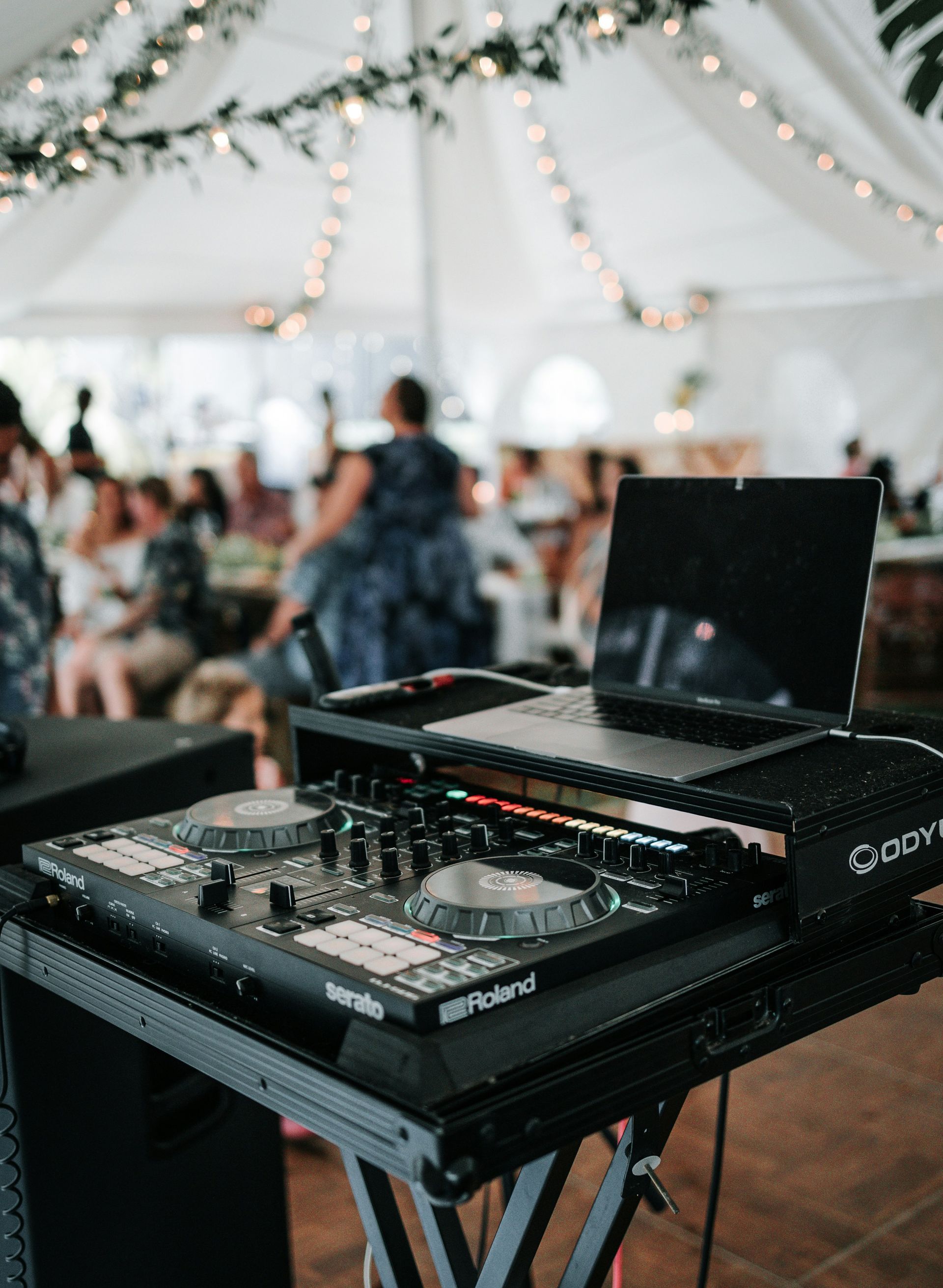 Attrezzatura da DJ con laptop e mixer durante un evento, con persone e tenda sullo sfondo.
