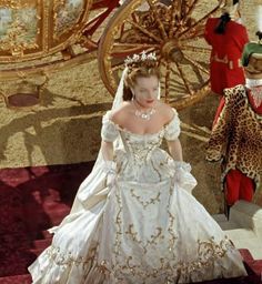 Una donna in un elaborato abito bianco scende le scale, con dettagli dorati, vicino a una carrozza e velluto rosso.