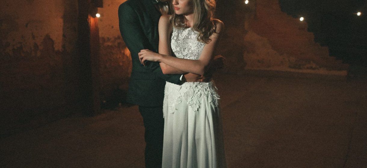 Una coppia si abbraccia in un ambiente rustico e scarsamente illuminato; la donna indossa un abito bianco, l'uomo un abito scuro.