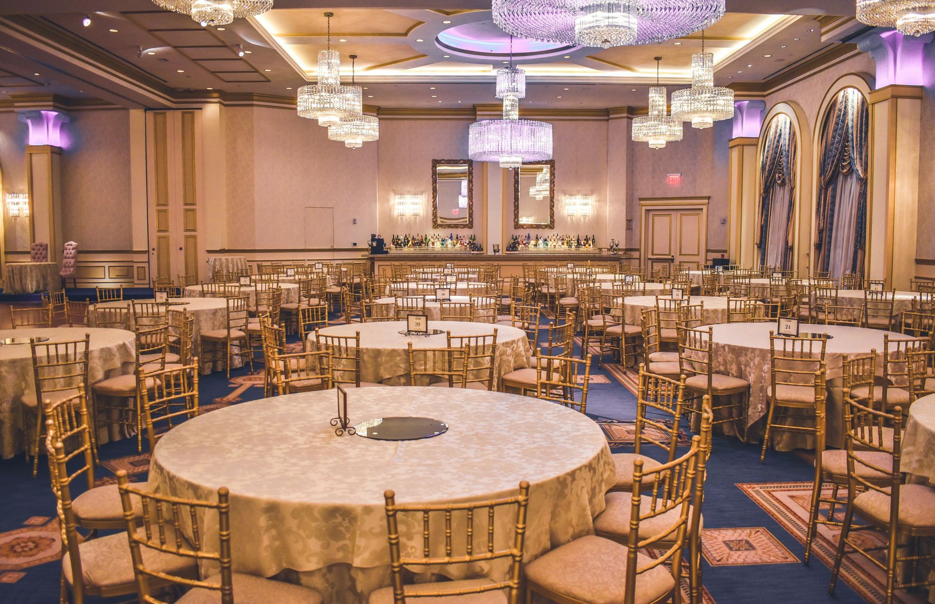 Sala da ballo formale con tavoli rotondi apparecchiati per un banchetto. Sedie e tovaglie dorate, lampadari e decorazioni murali.