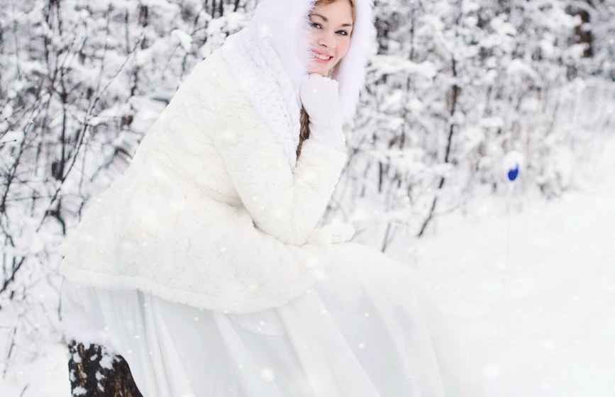 Una donna con camice bianco e cappuccio sorride in una foresta innevata.