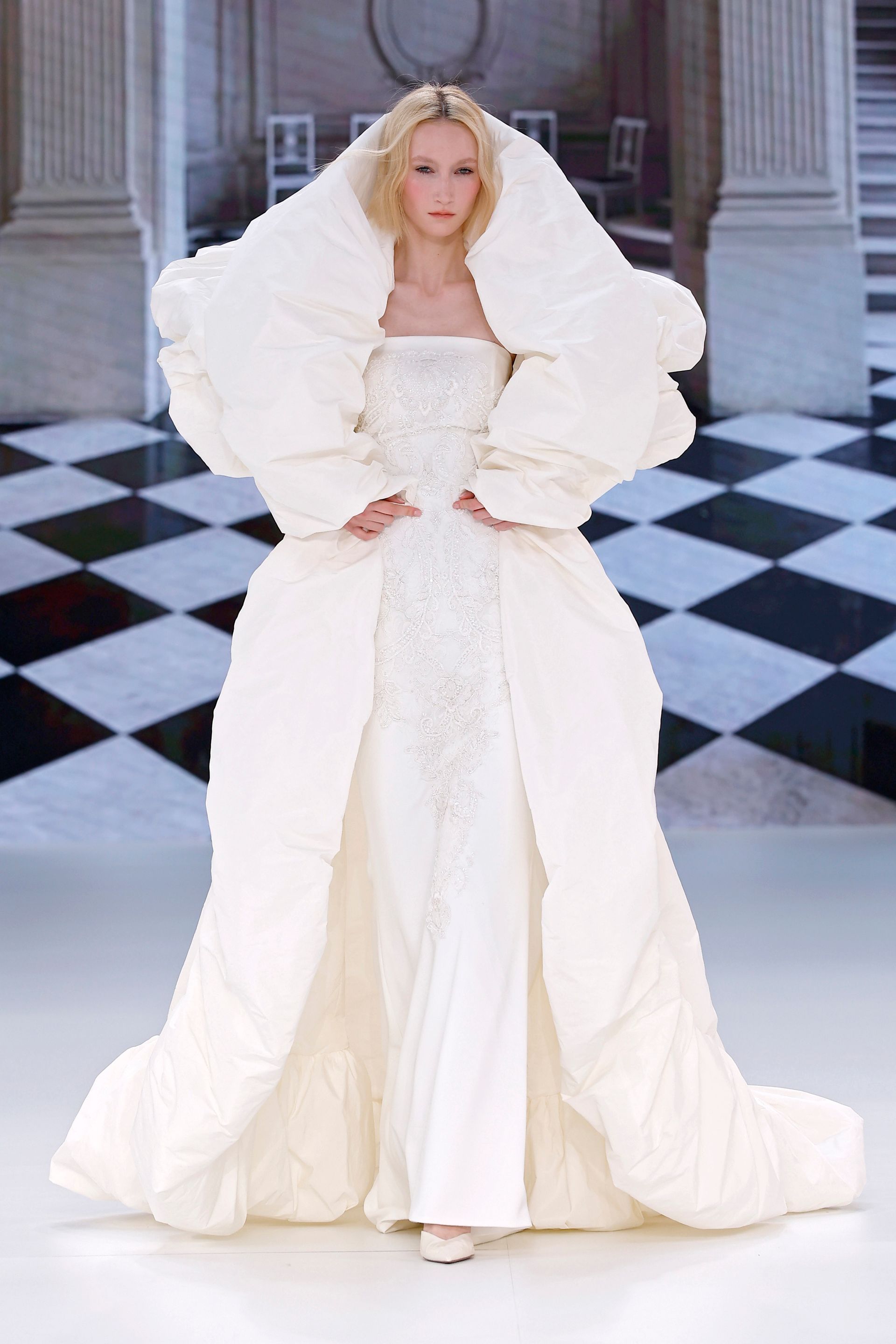 Donna in un abito bianco gonfio di alta moda con corpetto decorato con perline, su una passerella con pavimento a scacchi.