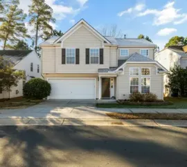 Suburban two-story beige house with white trim, garage, and driveway in front of trees under a blue sky